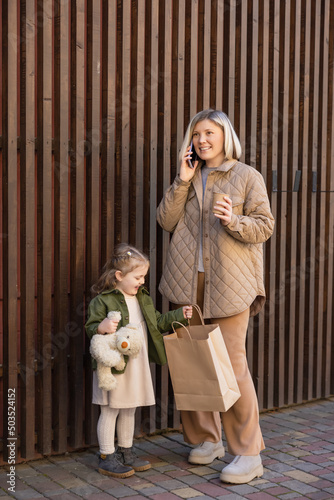 happy woman with paper cup talking on smartphone near daughter with teddy bear and shopping bag.