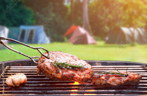 Roasted beef ripe steaks on flaming charcoal grill with blurred background of camping area in natural park