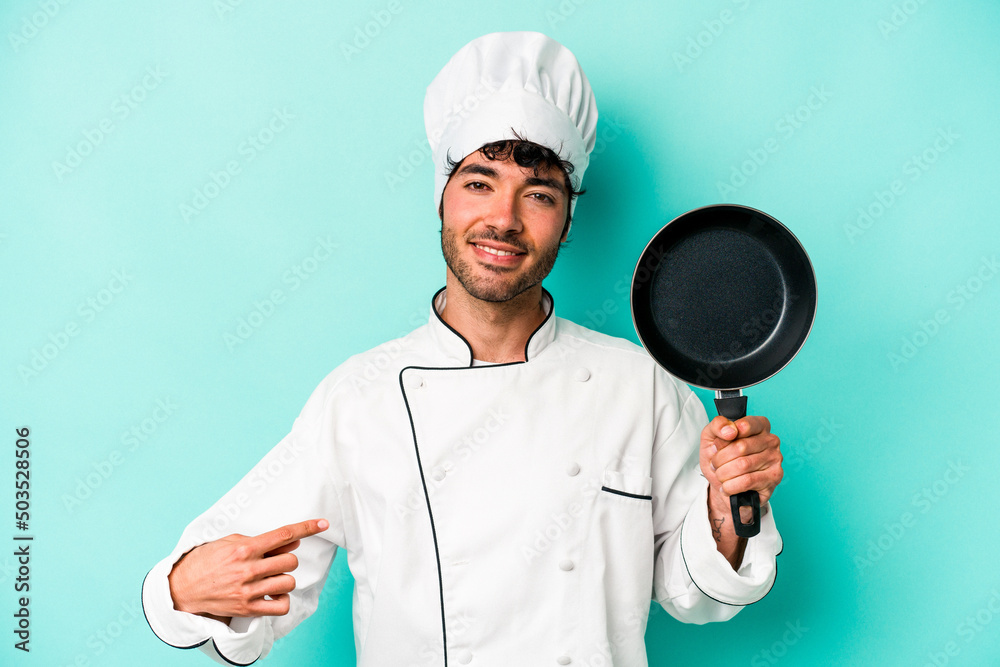 Young caucasian chef man holding flying pan isolated on blue background person pointing by hand to a shirt copy space, proud and confident Young caucasian chef man holding flying pan isolated on blue background person pointing by hand to a shirt copy space, proud and confident