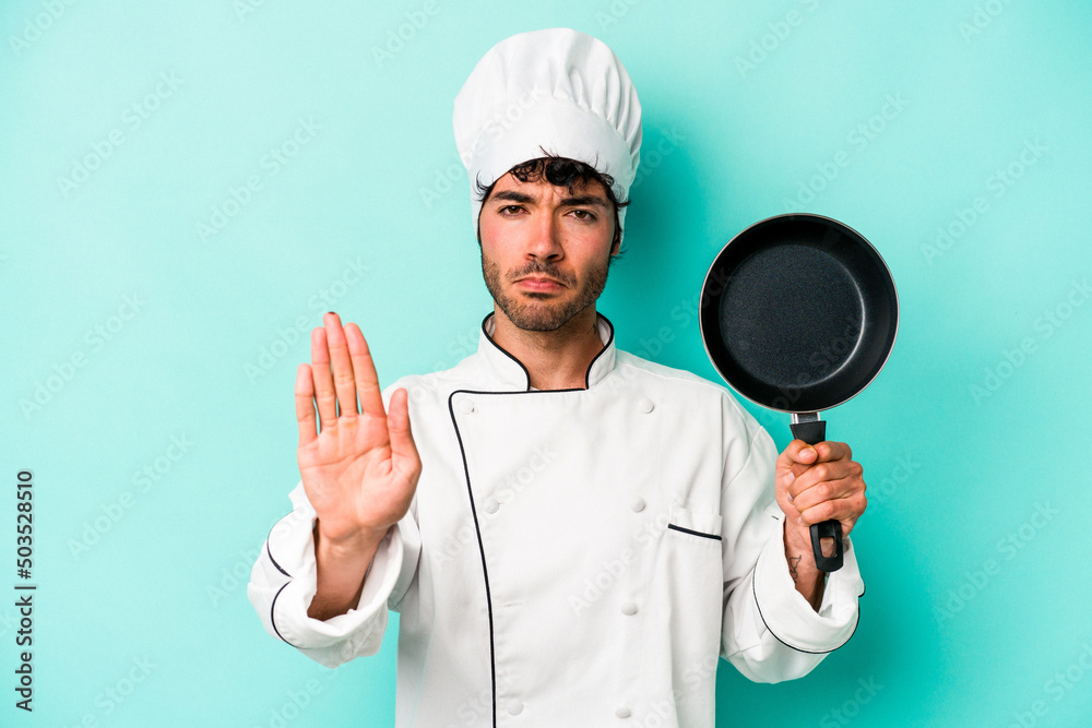 Young caucasian chef man holding flying pan isolated on blue background standing with outstretched hand showing stop sign, preventing you. Young caucasian chef man holding flying pan isolated on blue background standing with outstretched hand showing stop sign, preventing you.