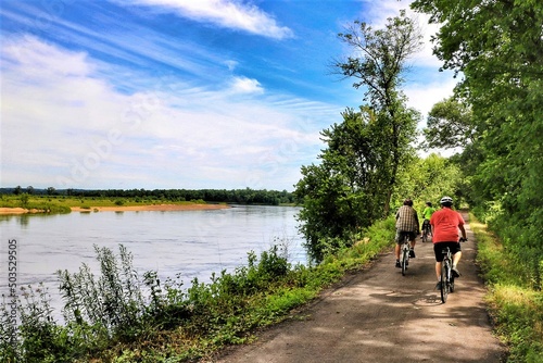Beneath a blue sky with white clouds on a summer day in Wisconsin, three cyclists on a dirt path pass through a forested landscape and alongside a peaceful, reflective river with a sandy beach on the 