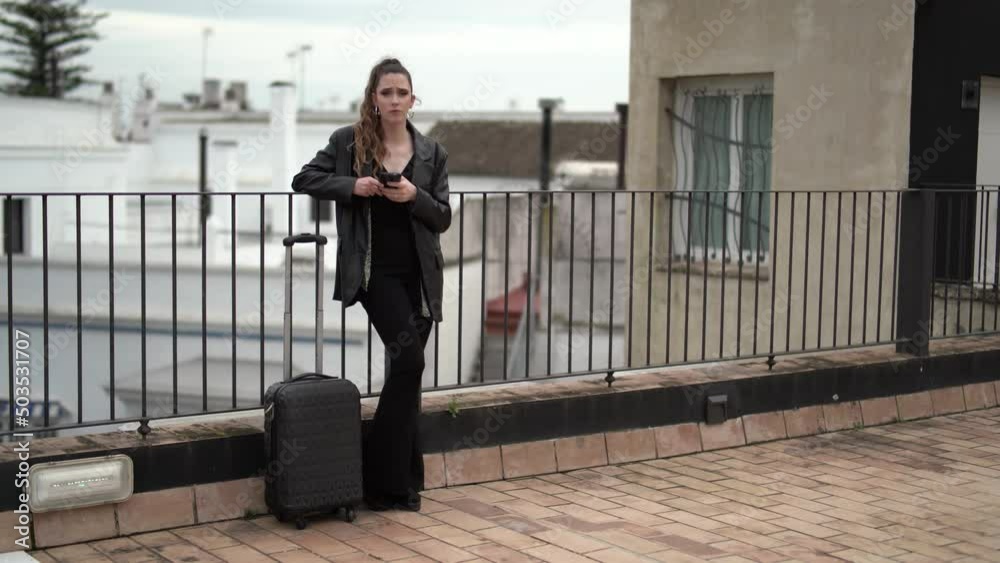 Chica joven delgada con pantalon y blusa negra con maleta de viaje en terraza de hotel Chica joven delgada con pantalon y blusa negra con maleta de viaje en terraza de hotel