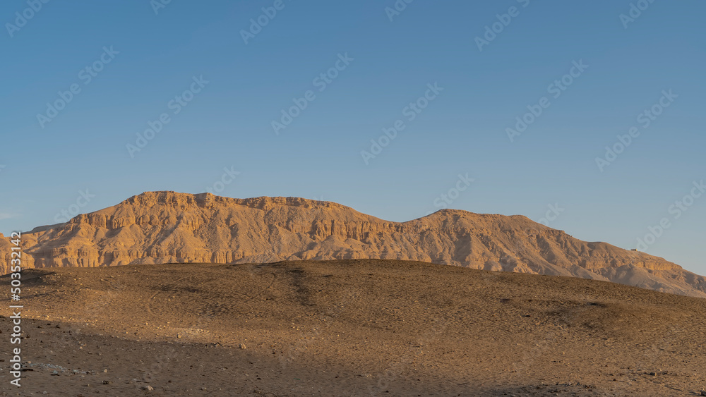 A picturesque rock devoid of vegetation against a clear blue sky. In the foreground is a desert sand dune. Egypt. Luxor.