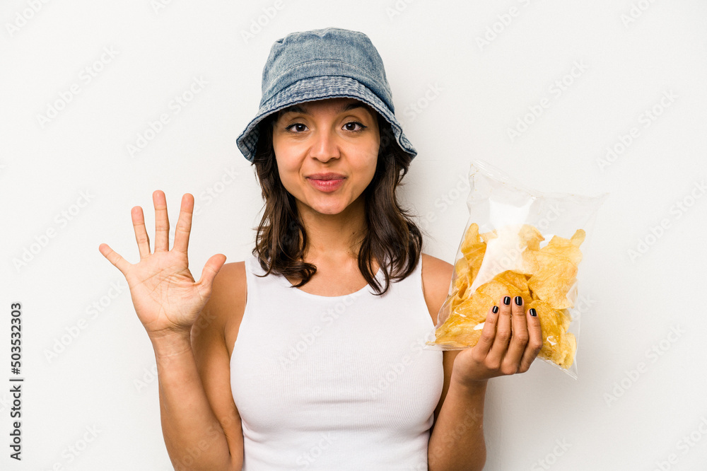 Young hispanic woman holding a bag of chips isolated on white background smiling cheerful showing number five with fingers. Young hispanic woman holding a bag of chips isolated on white background smiling cheerful showing number five with fingers.