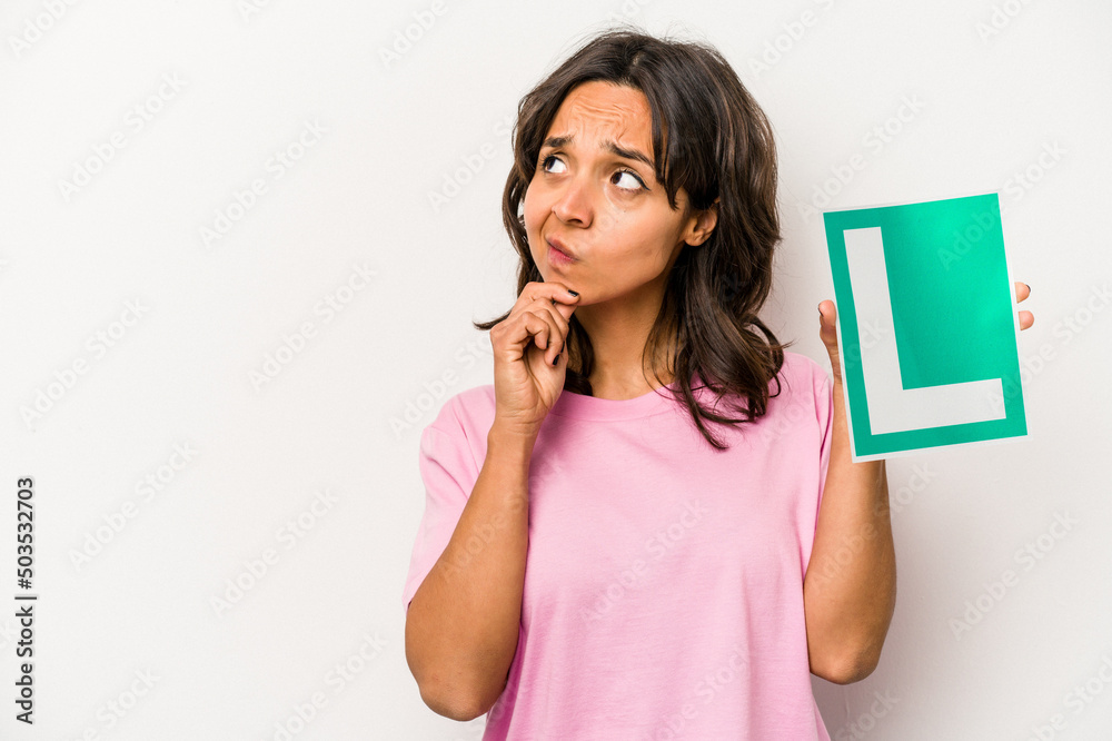 Young hispanic woman holding a beginner driver sign isolated on white background looking sideways with doubtful and skeptical expression. Young hispanic woman holding a beginner driver sign isolated on white background looking sideways with doubtful and skeptical expression.