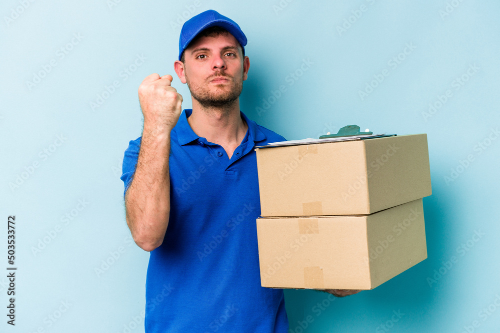 Young caucasian delivery man isolated on blue background showing fist to camera, aggressive facial expression. Young caucasian delivery man isolated on blue background showing fist to camera, aggressive facial expression.
