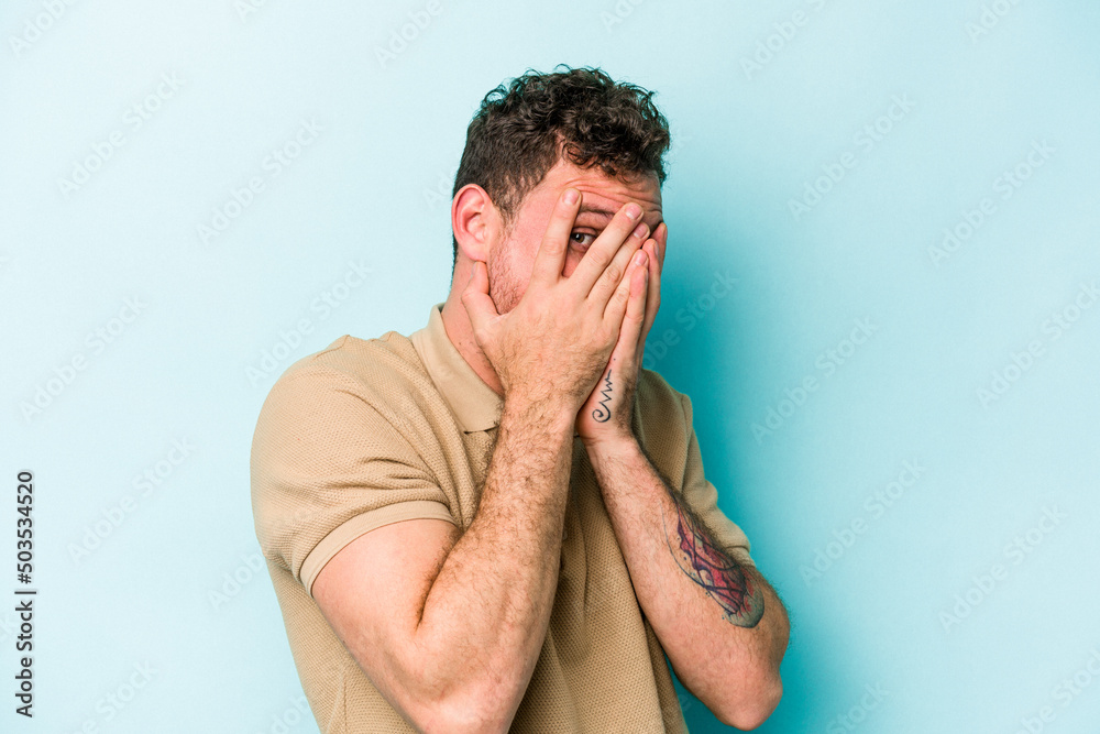 Young caucasian man isolated on blue background blink through fingers frightened and nervous. Young caucasian man isolated on blue background blink through fingers frightened and nervous.