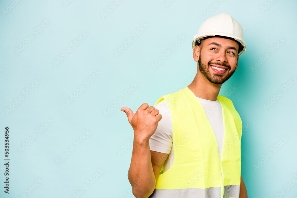 Young laborer hispanic man isolated on blue background points with thumb finger away, laughing and carefree. Young laborer hispanic man isolated on blue background points with thumb finger away, laughing and carefree.