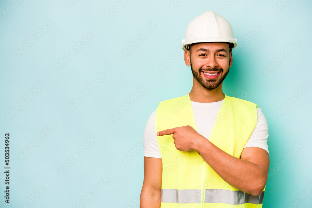 Young laborer hispanic man isolated on blue background smiling and pointing aside, showing something at blank space.