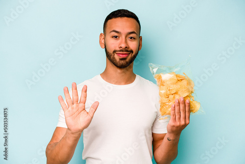 Young hispanic man holding a bag of chips isolated on blue background smiling cheerful showing number five with fingers.