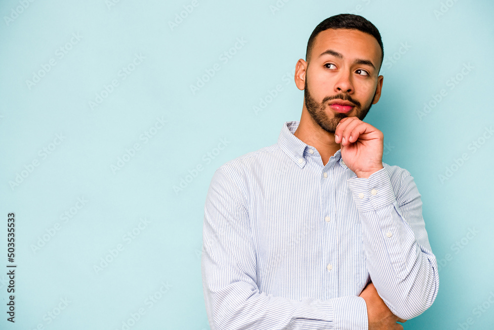 Young hispanic man isolated on blue background looking sideways with doubtful and skeptical expression. - obrazy, fototapety, plakaty 