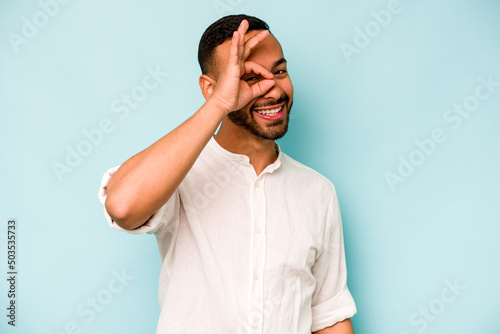Young hispanic man isolated on blue background excited keeping ok gesture on eye. © Asier