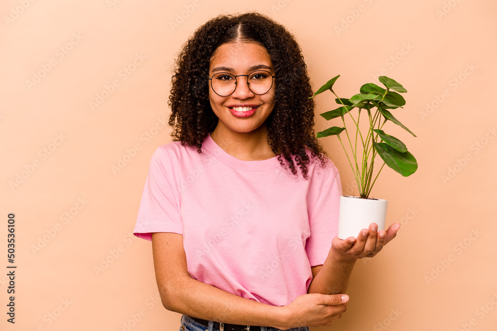 Young African American woman holding a plant isolated on beige background laughing and having fun. Young African American woman holding a plant isolated on beige background laughing and having fun.