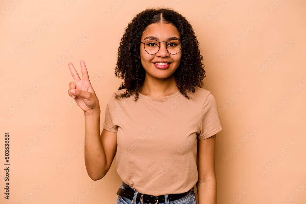Young African American woman isolated on beige background joyful and carefree showing a peace symbol with fingers. Young African American woman isolated on beige background joyful and carefree showing a peace symbol with fingers.