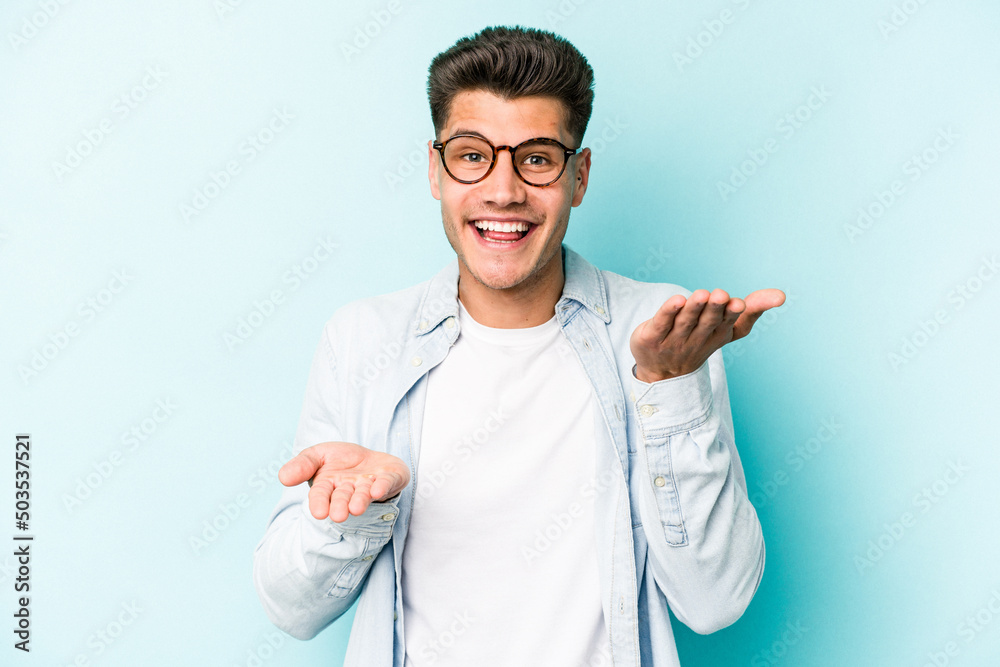 Young caucasian man isolated on blue background makes scale with arms, feels happy and confident. Young caucasian man isolated on blue background makes scale with arms, feels happy and confident.