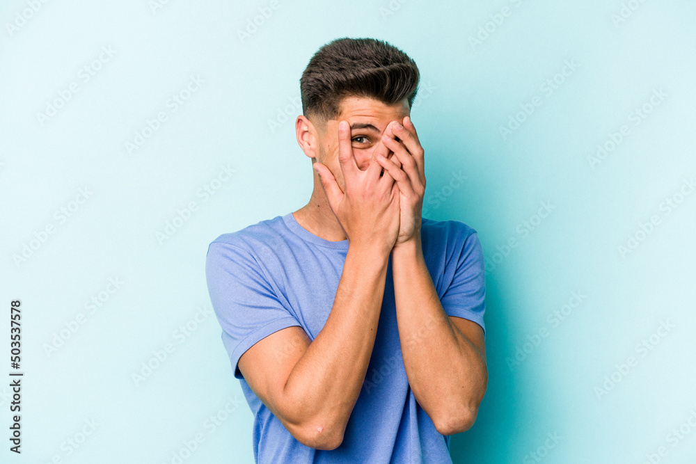 Young caucasian man isolated on blue background blink at the camera through fingers, embarrassed covering face.