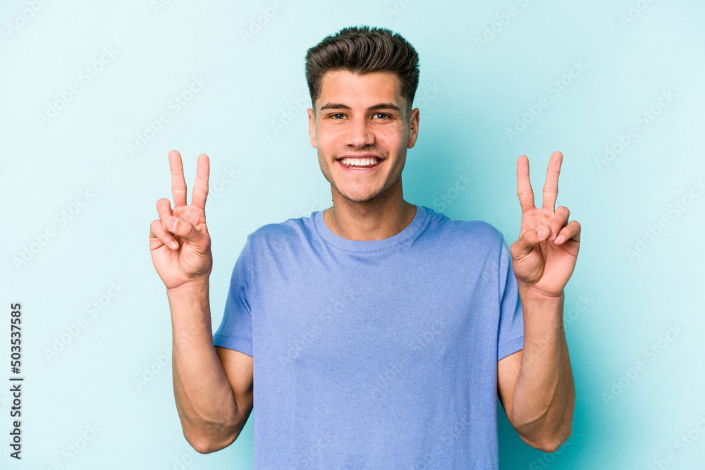 Young caucasian man isolated on blue background showing victory sign and smiling broadly. Young caucasian man isolated on blue background showing victory sign and smiling broadly.