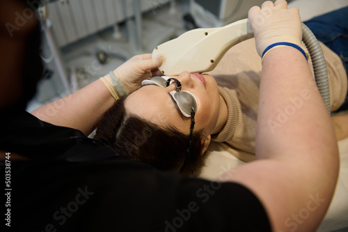 Young pretty woman wearing UV protective goggles, lying down on daybed while receiving hair removal laser procedure on her face in a wellness spa clinic