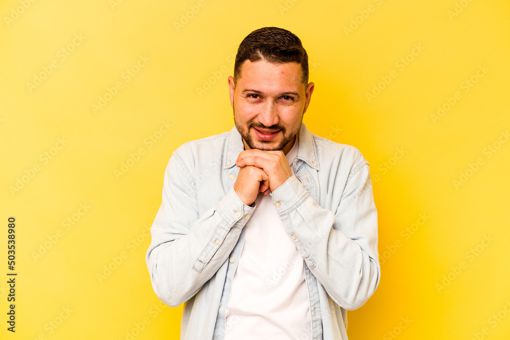 Young hispanic man isolated on yellow background keeps hands under chin, is looking happily aside. Young hispanic man isolated on yellow background keeps hands under chin, is looking happily aside.