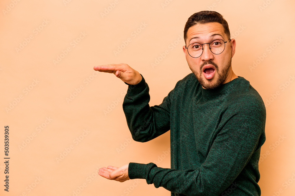 Young hispanic man isolated on beige background shocked and amazed holding a copy space between hands. Young hispanic man isolated on beige background shocked and amazed holding a copy space between hands.