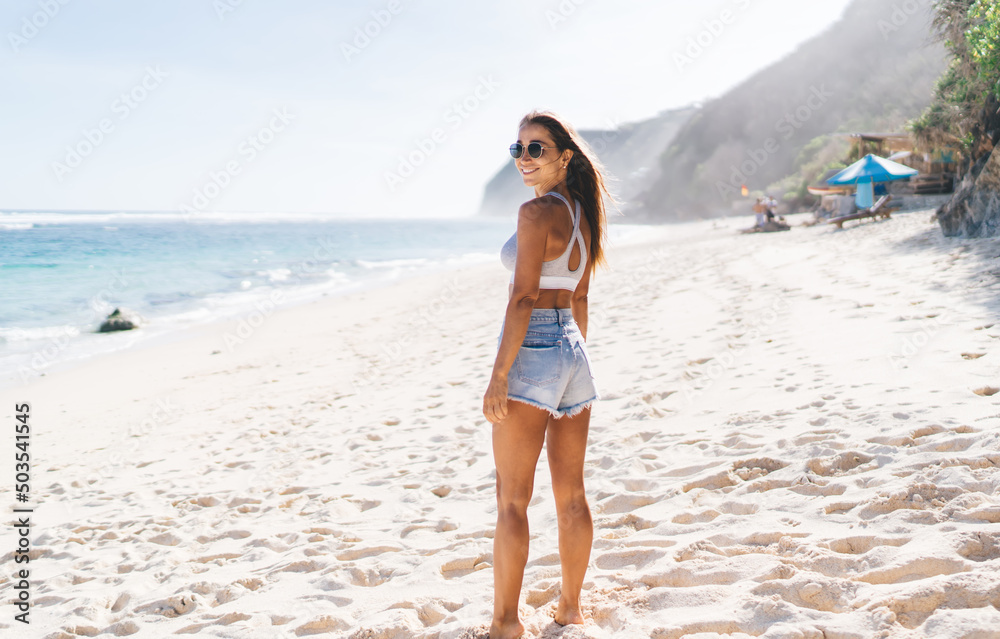 Full length portrait of cheerful female smiling at camera during touristic journey at Sri Lanka, happy Caucasian woman in sunglasses enjoying travel vacations posing at seashore with white sand