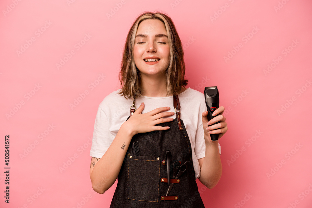 Young hairdresser woman holding scissors isolated on pink background laughs out loudly keeping hand on chest. Young hairdresser woman holding scissors isolated on pink background laughs out loudly keeping hand on chest.