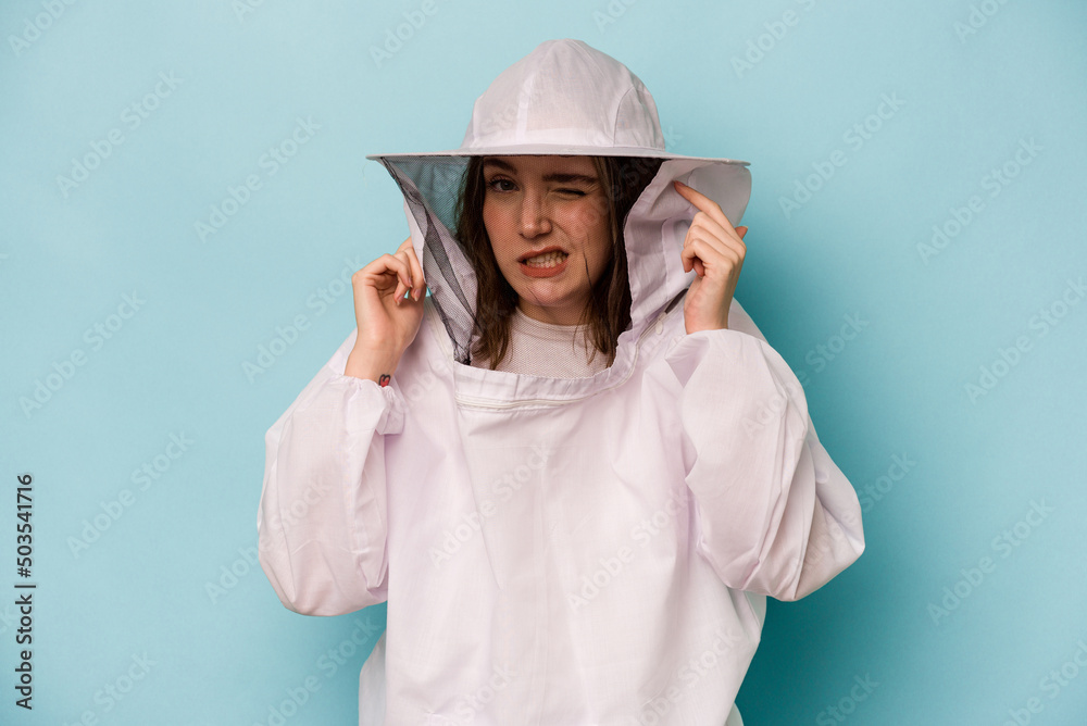 Young caucasian beekeeper woman isolated on blue background covering ears with hands. Young caucasian beekeeper woman isolated on blue background covering ears with hands.