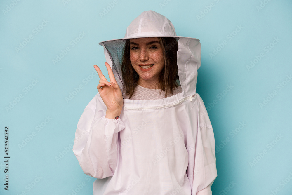 Young caucasian beekeeper woman isolated on blue background joyful and carefree showing a peace symbol with fingers. Young caucasian beekeeper woman isolated on blue background joyful and carefree showing a peace symbol with fingers.