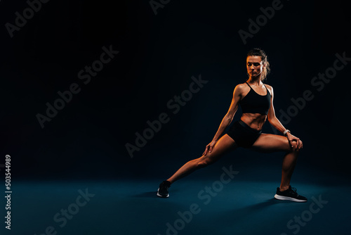 Young healthy woman in fitness wear warming up her body before training on black background