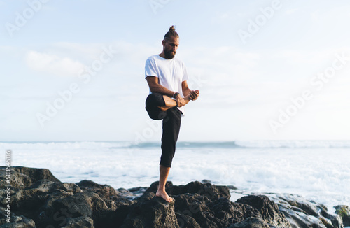 Caucaisan man in casual sportswear doing balance asana with raising leg breathing at coastline  calm male keeping fit and healthy lifestyle with yoga practice and stretching exercises at seashore