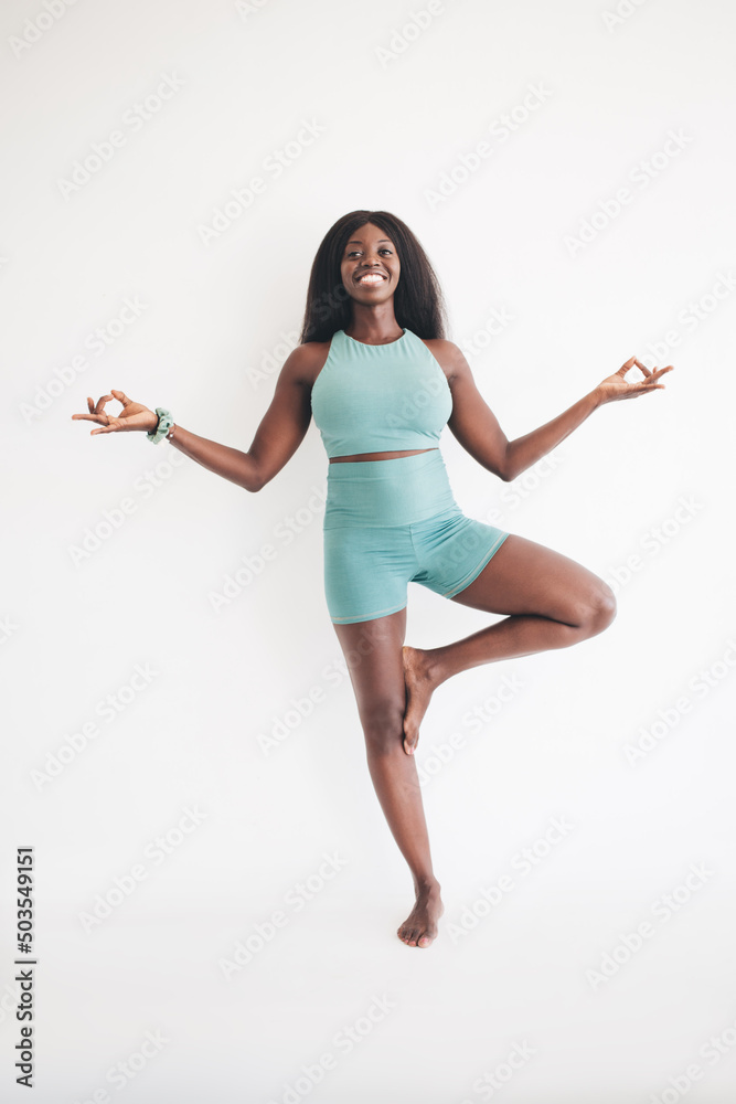 Cheerful African American woman in sportive tracksuit practicing balance pose in tree asana during yoga in white aerobic studio, happy dark skinned fit girl enjoying body positive and vitality