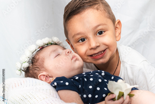 latin boy with a big smile, posing for the picture next to his baby sister, the baby is lying on a white pillow, with her eyes closed, a crown of white roses and holding a rose. photo