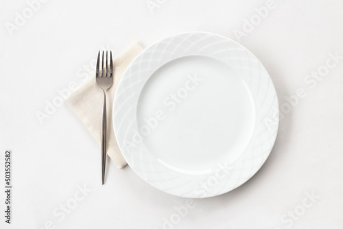 Empty white plate with fabric napkin and a fork on white table. Top view.