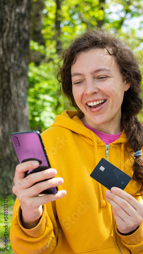 Excited young woman customer or buyer shopping online on mobile with credit card, wearing yellow hoodie on green garden background. Making internet payment, online shopping, secure banking.Vertical photo