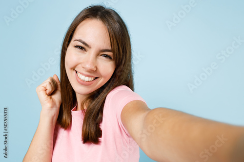 Close up young smiling happy woman 20s wear pink t-shirt doing selfie shot pov on mobile cell phone do winner gesture isolated on pastel plain light blue background studio. People lifestyle concept.