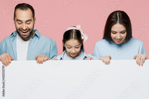 Young fun parents mom dad with child kid daughter teen girl in blue clothes hold empty billboard for promotional content, area place for text or image isolated on plain pastel light pink background.