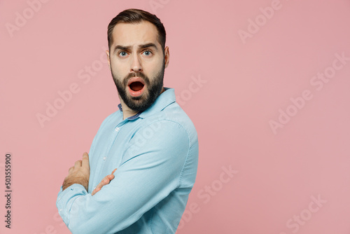 Side view young strict sad indignant man 20s wear classic blue shirt look camera holding hands crossed folded isolated on plain pastel light pink background studio portrait. People lifestyle concept.