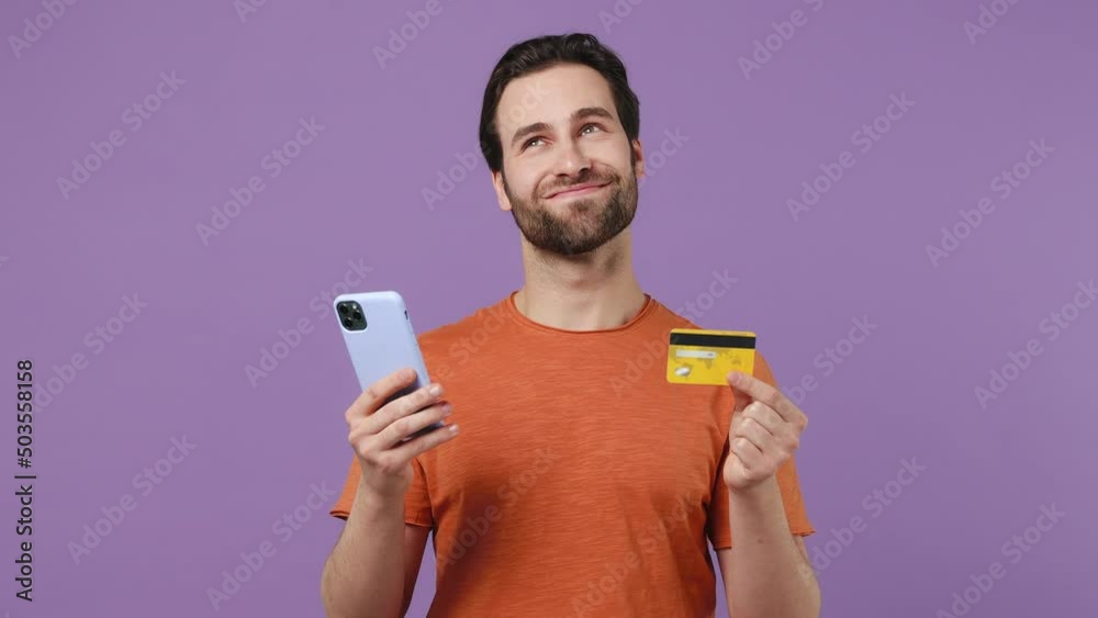 Dreamful happy young brunet bearded man 20s in red t-shirt using mobile cell phone hold in hand credit bank card doing online shopping isolated on plain pastel light purple background studio portrait