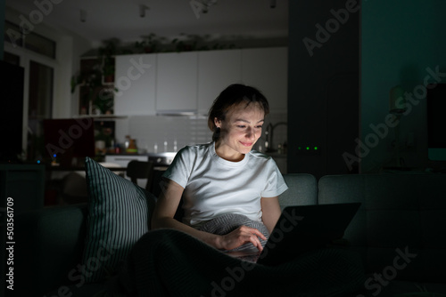 Smiling girl looking at computer screen, talking in video chat. Cheerful woman freelancer sitting on couch, having video call with friends at night at home. Female working on laptop in dark room. 