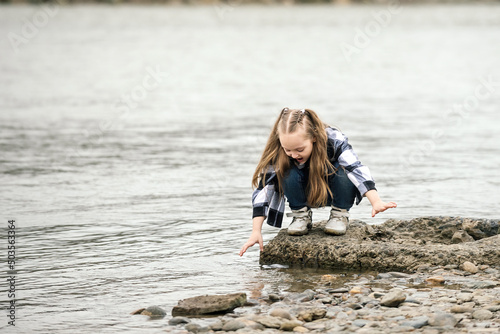 Fototapeta Naklejka Na Ścianę i Meble -  a girl with blond long hair, in a fashionable plaid shirt and jeans, has fun by the river on a walk during school holidays