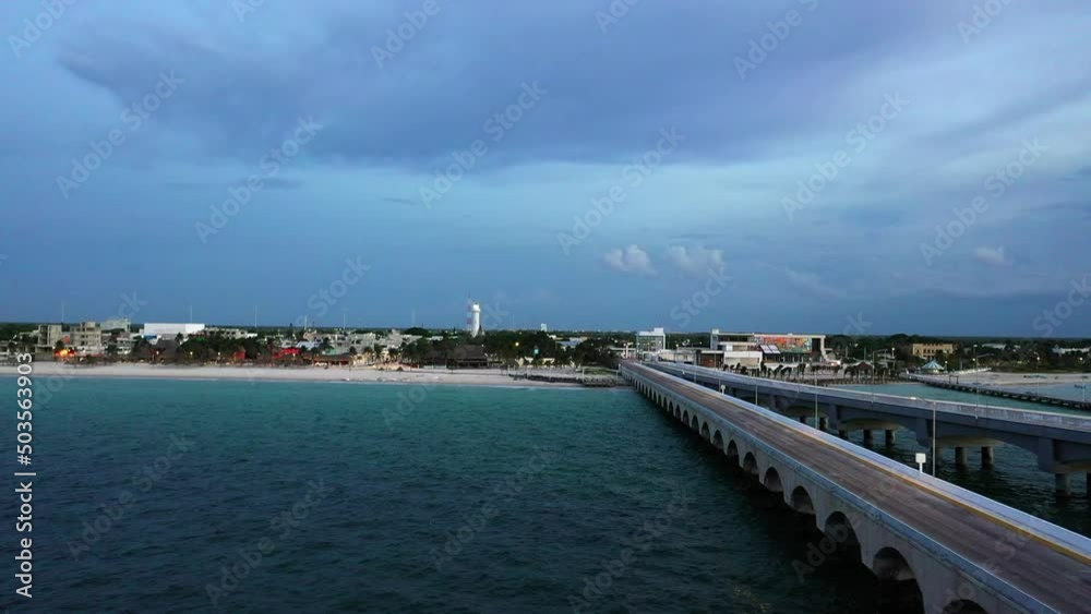 Flying along the lanes of a long pier, facing a lighthouse and the beach, as the sun sets. The port of a small coastal town. Flying along the lanes of a long pier, facing a lighthouse and the beach, as the sun sets. The port of a small coastal town.