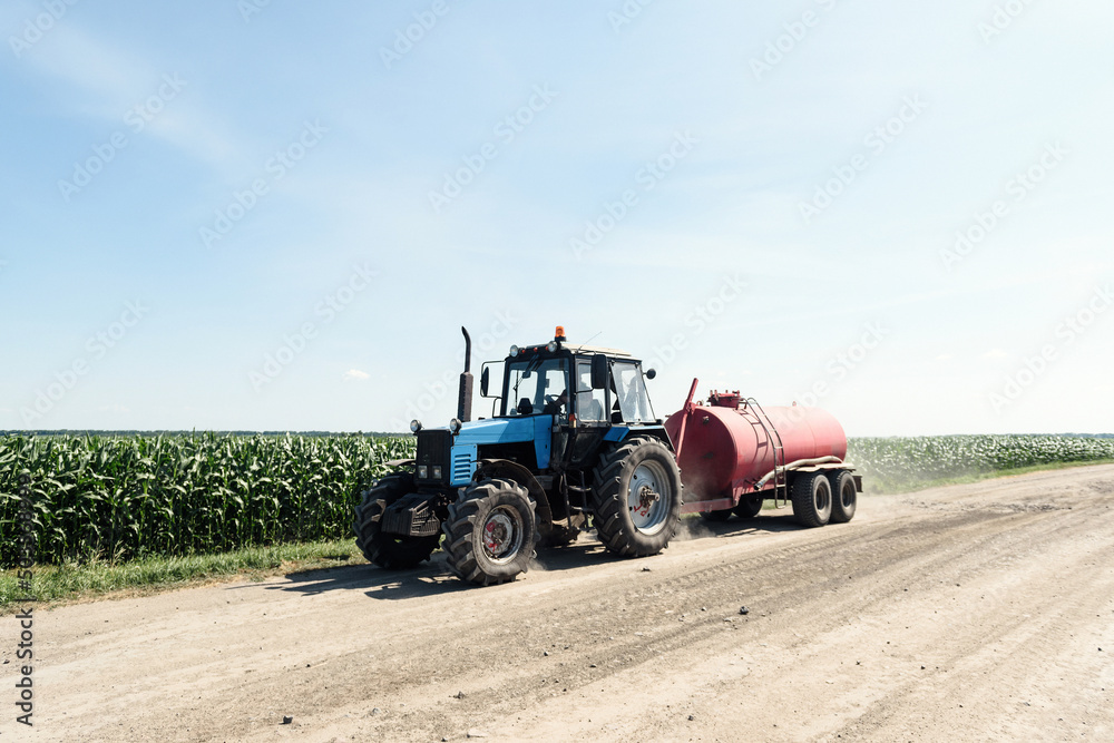 Obraz premium Tractor carries a barrel of water on the field near the corn on a background of sky
