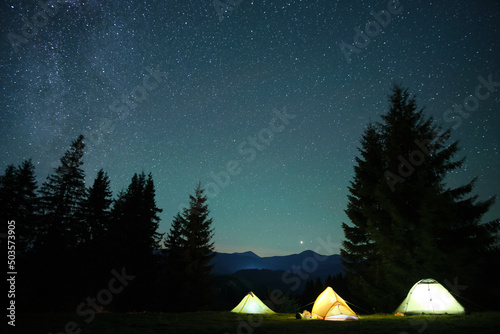 Brightly illuminated camping tents glowing on campsite in dark mountains under night stars covered sky. Active lifestyle and traveling concept