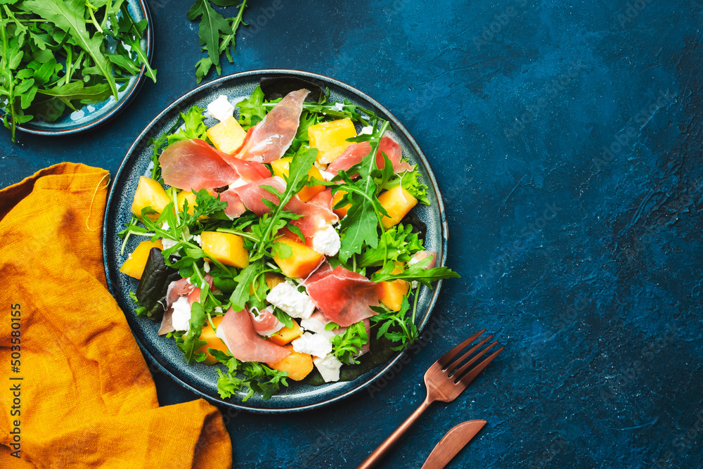 Delicious summer fresh salad with cantaloupe melon, prosciutto, soft cheese and arugula on blue table background, top view, copy space Delicious summer fresh salad with cantaloupe melon, prosciutto, soft cheese and arugula on blue table background, top view, copy space