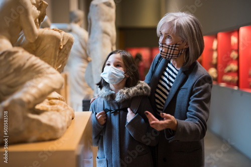 Portrait of positive intelligent senior woman and cute interested preteen girl wearing protective masks viewing ancient sculptures in museum. Forced precautions in pandemic