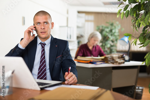 Portrait of focused busy entrepreneur solving business issues by phone while sitting at office table with laptop