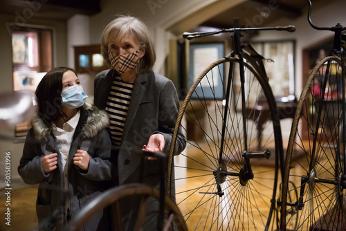 Curious tween schoolgirl and friendly elderly female tutor in face masks viewing vintage bicycle in museum of history of technology with interest. Knowledge concept. New normal in coronavirus pandemic