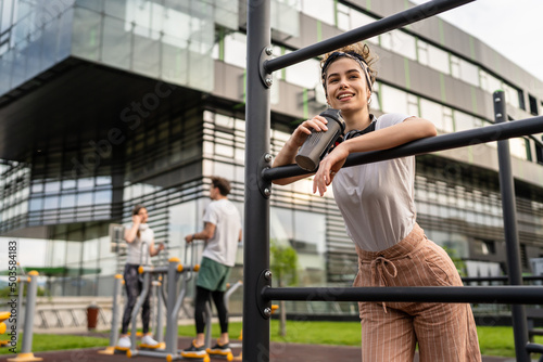 One caucasian woman taking a brake during outdoor training in the park outdoor gym resting on the bars with supplement shaker in hand drinking water or supplementation happy smile copy space