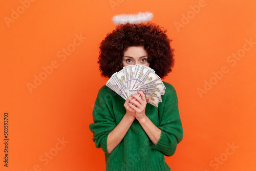 Portrait of angelic woman with Afro hairstyle wearing green casual style sweater and nimb covering half of face with big fan of dollars banknotes. Indoor studio shot isolated on orange background.