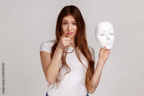 Portrait of dark haired woman pointing finger on you, holding white mask with unknown face, blaming you in duplicity, wearing white T-shirt. Indoor studio shot isolated on gray background. photo